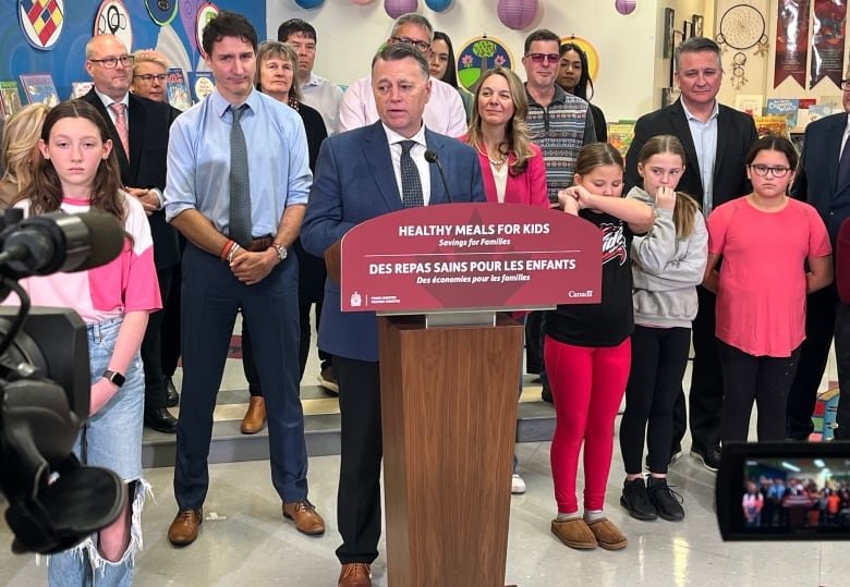 A man in a suit stands at a podium speaking, with several other people gathered behind him during a news conference at a school.