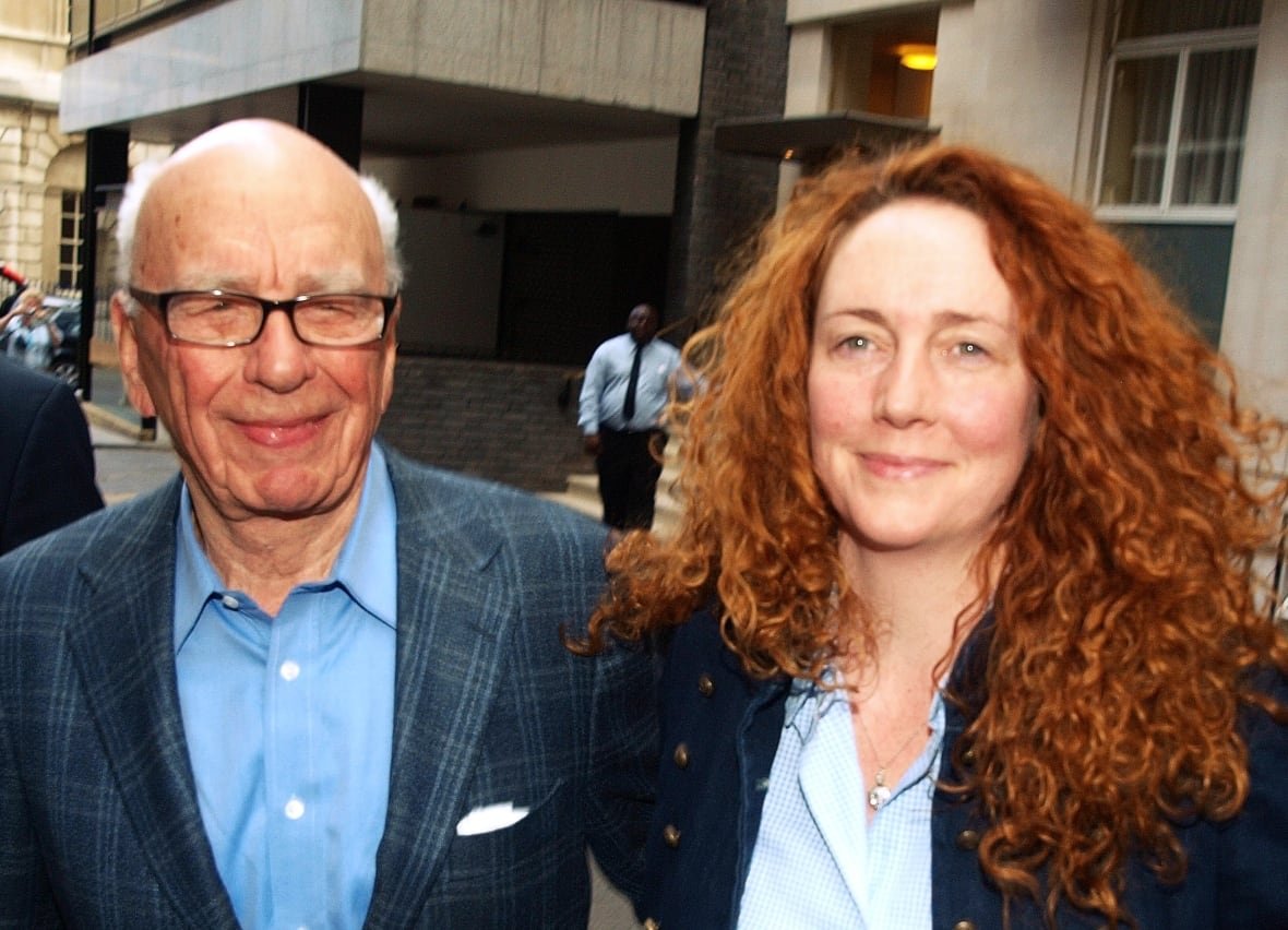 A bald older man in glasses wearing a checked blazer and collared shirt stands next to a woman with curly red hair past her shoulders.
