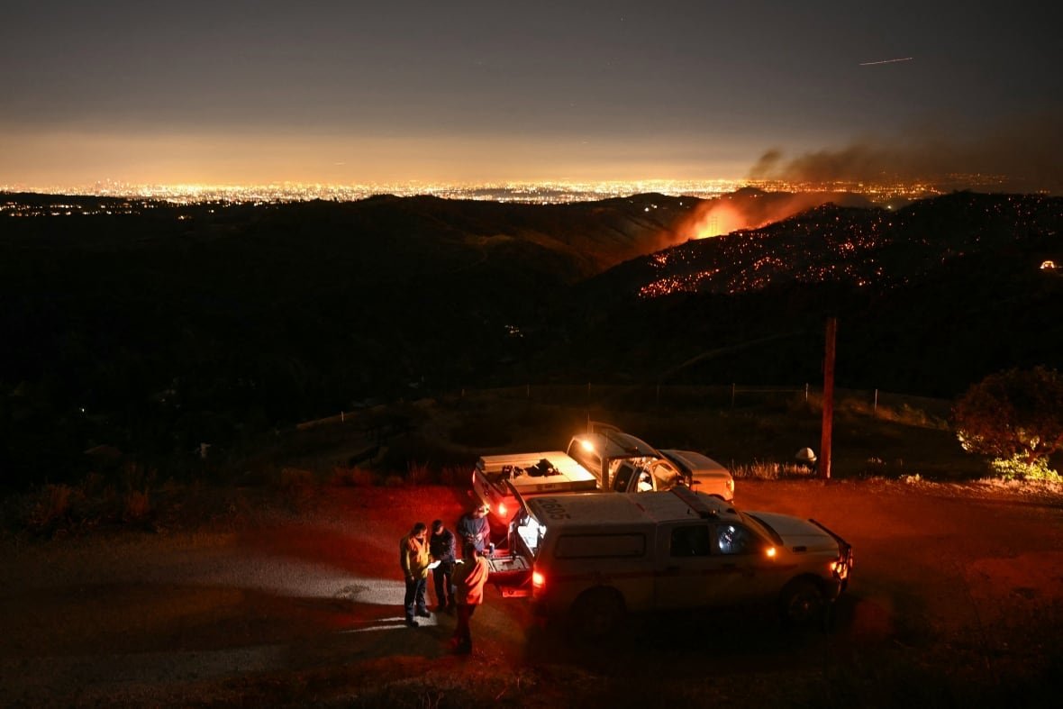 Firefighters gather near vehicles as a wildfire burns on a hill in the background.