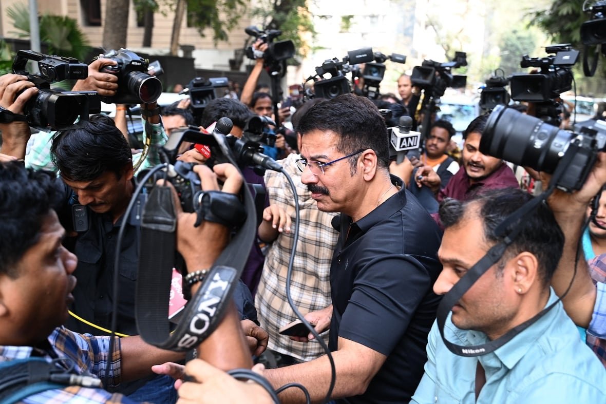 A crowd of photographers hold their cameras above their heads. They are pointed at a man in the middle of the crowd who appears to be walking through them.