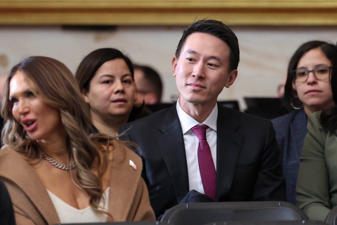 A man in a business suit sits in an audience among three women. He is looking at something to his right.