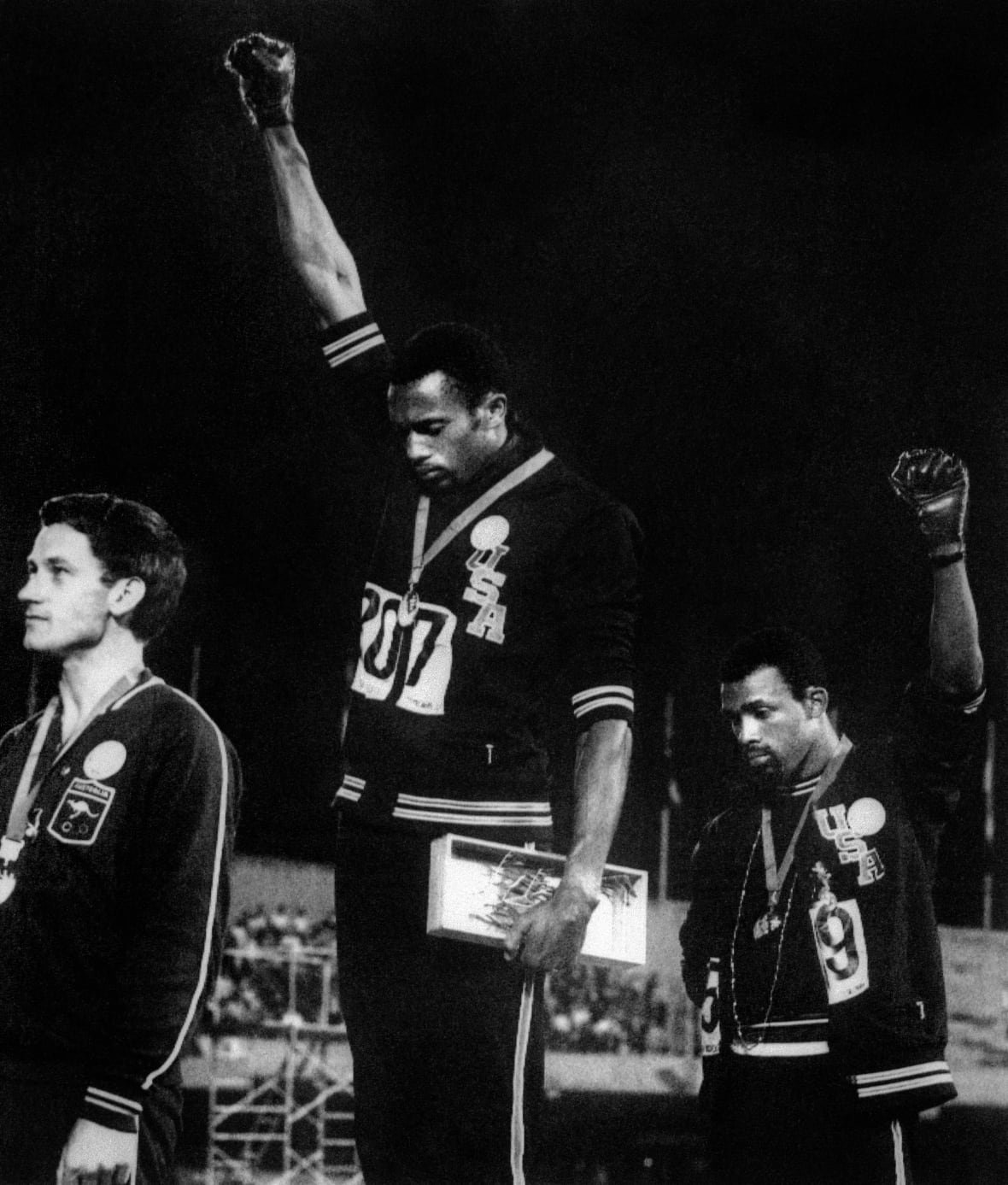 Three men stand on an Olympic podium, the two men at right raise their fists in a salute.