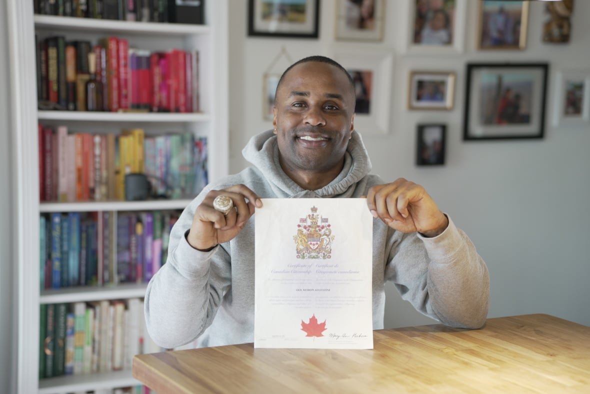 akil-augustine A smiling man wearing a hoodie and a large NBA championship ring on his right hand sits at a wooden table in front of a bookshelf holding a document with the Canadian coat of arms at the top and a large maple leaf at the bottom.
