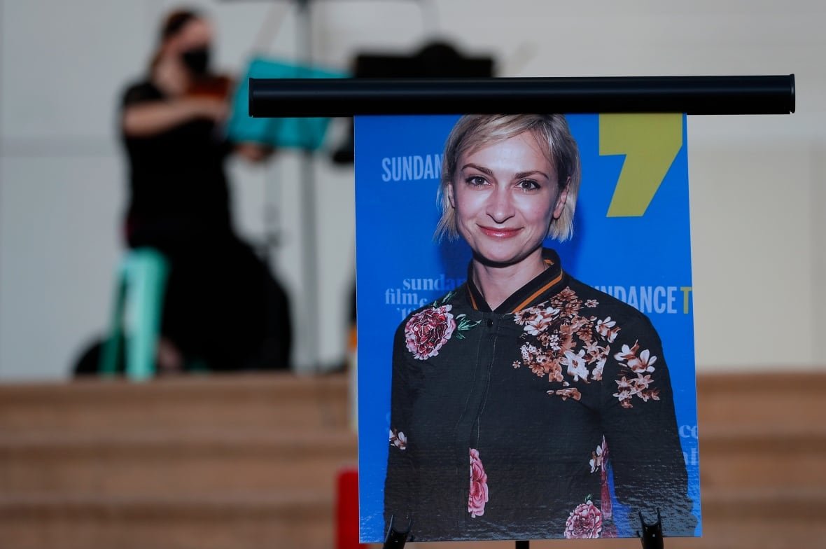 A large picture of a smiling blonde woman is propped up on an easel for display. In the blurred distance, a seated person plays a violin.