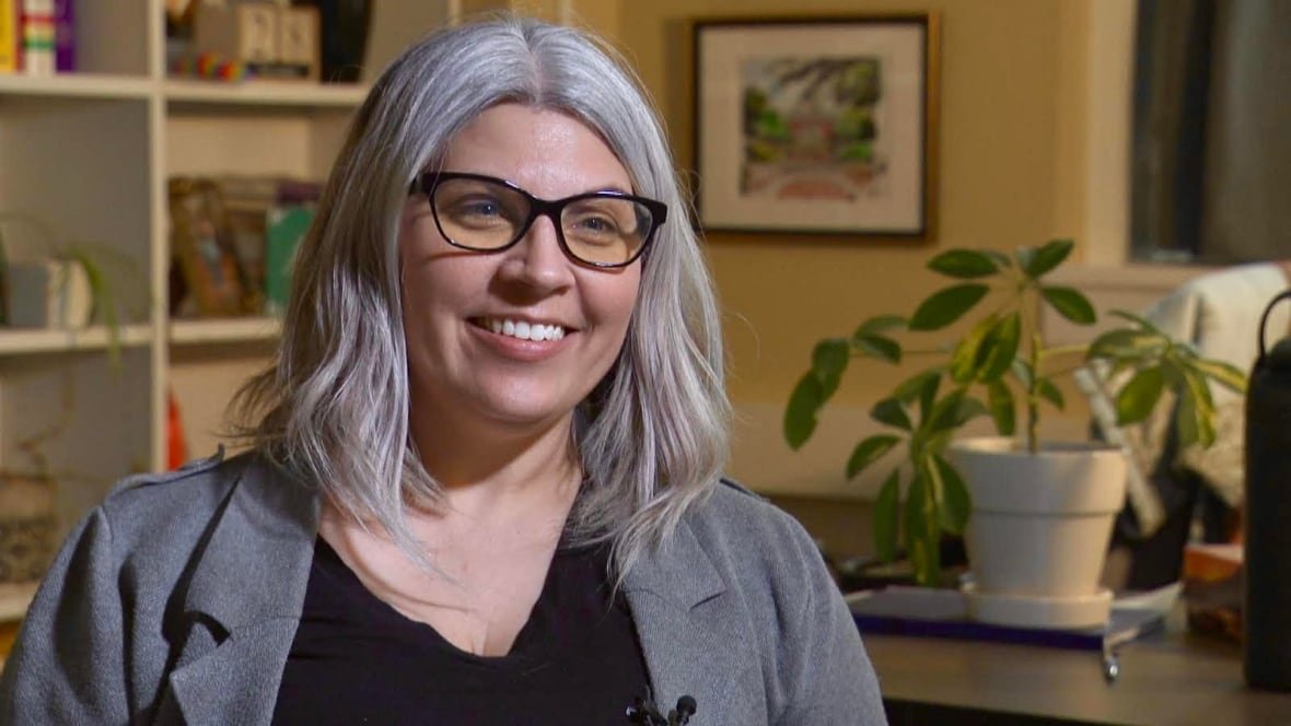 amanda-manning A young woman with grey hair, black rimming glasses and a grey blazer, smiles at the camera with pearly white teeth.