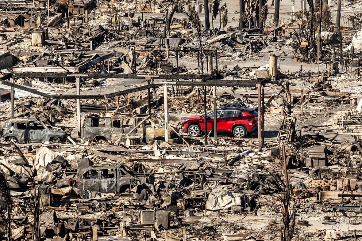 A bright red car drives past destroyed and burnt buildings and vehicles.