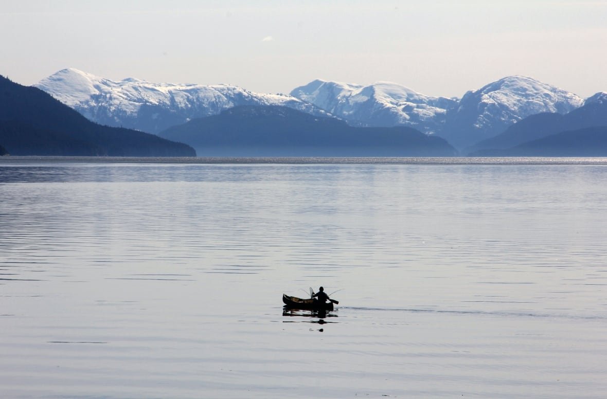 A long shot of a lone fisherman on a vast lake with snow-capped mountains in the distance.