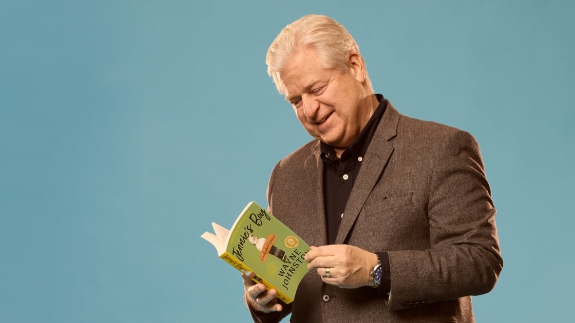 A white man with white hair flips through a green book wearing a brown sports jacket. He is in front of a blue background.