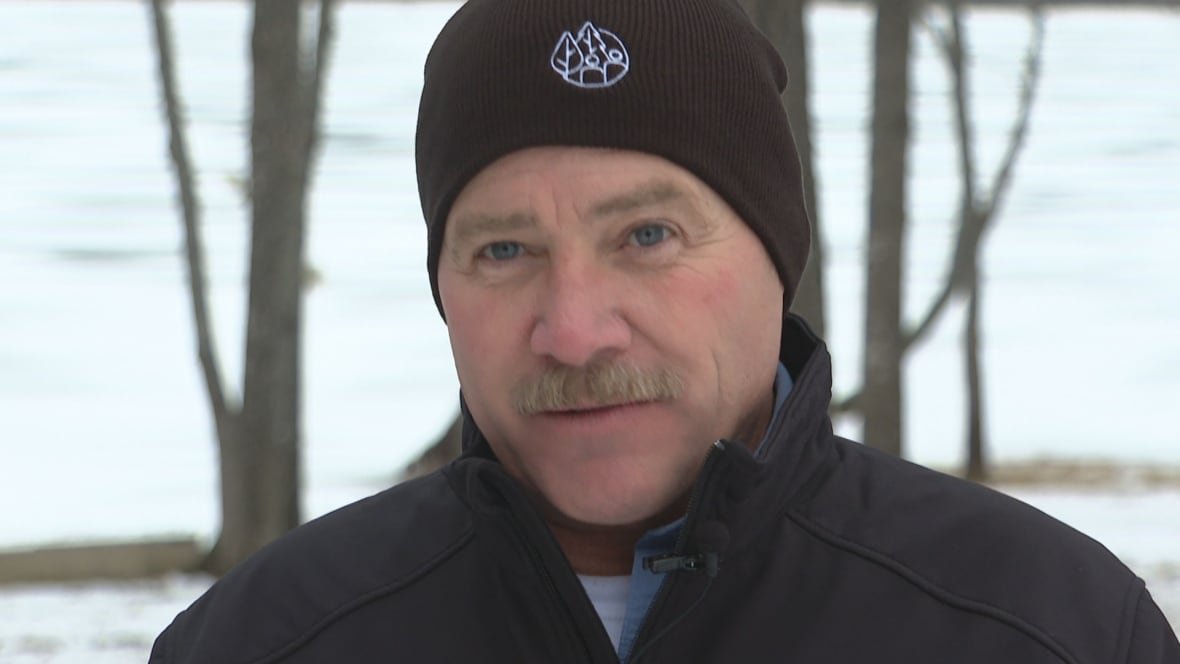Man with mustache in black toque and jacket stands in front of trees and icy lake.