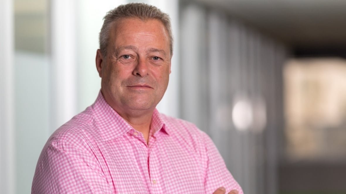 cybersecurity-expert-tony-anscombe A man in a pink-and-white checked dress shirt smiles at the camera while standing in an indoor hallway seen out of focus behind him.