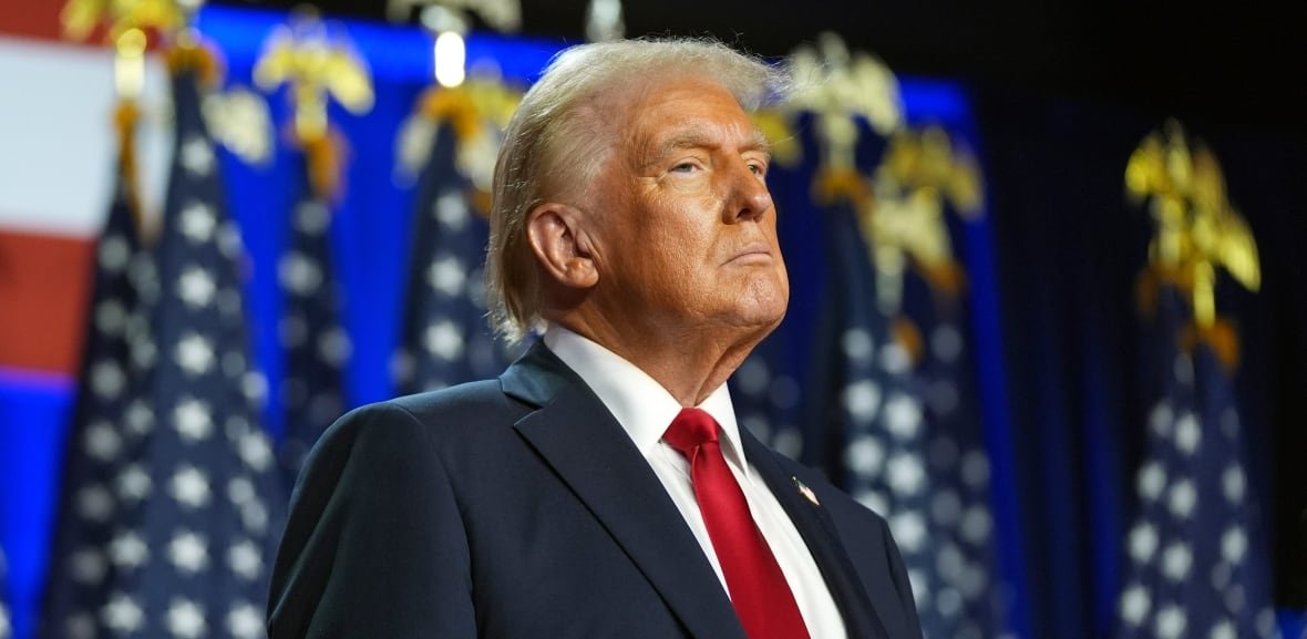 A man in a dark suit and red tie is seen on stage in front of several U.S. flags.