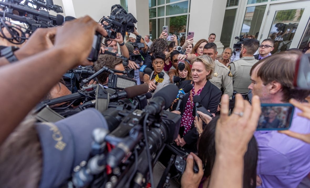 A woman stands in the centre of a tightly-packed crowd of people, speaking into a microphone. In front of her, everyone is holding a camera or microphone pointed at her, while behind her stands some police officers or security.