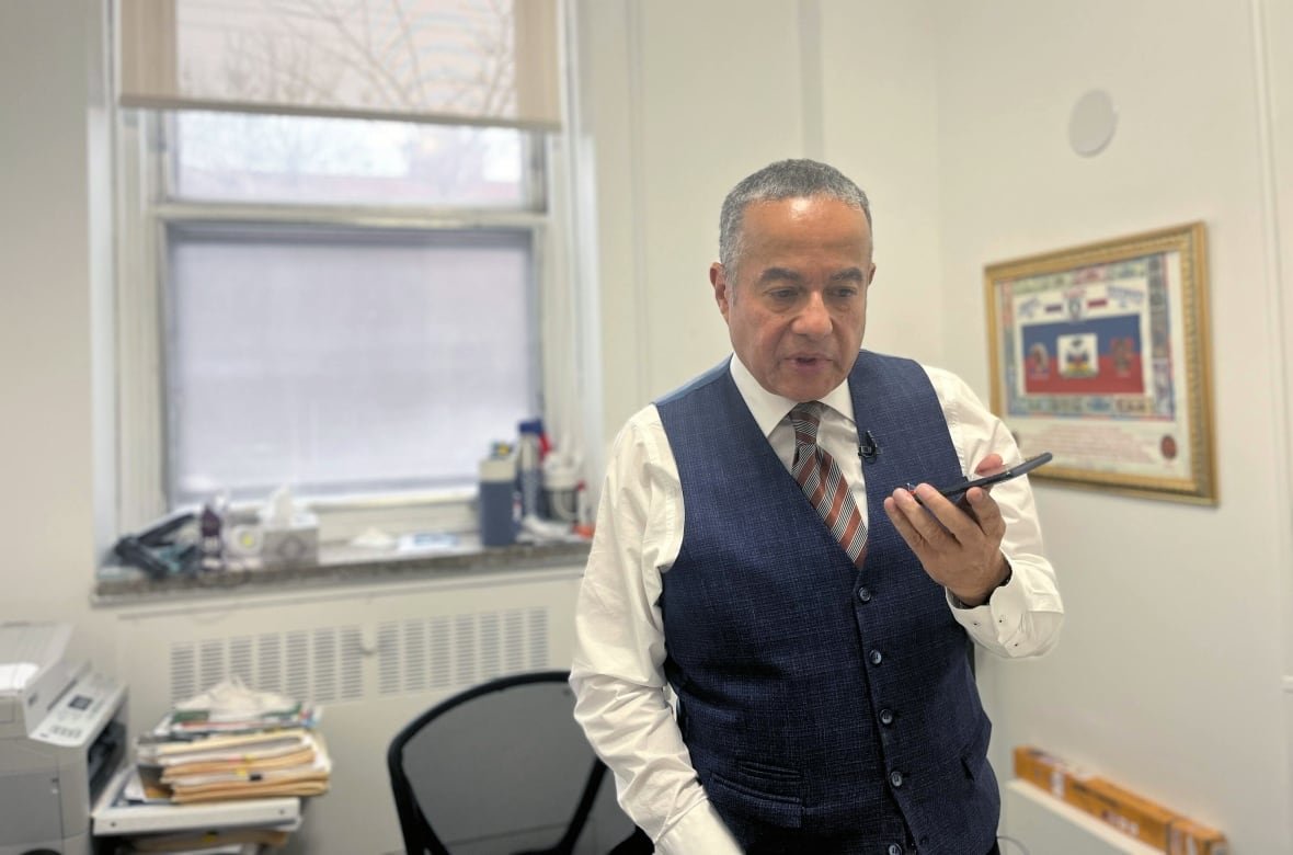 A man in a vest speaks into a phone inside a white office, with a Haitian flag on the wall.