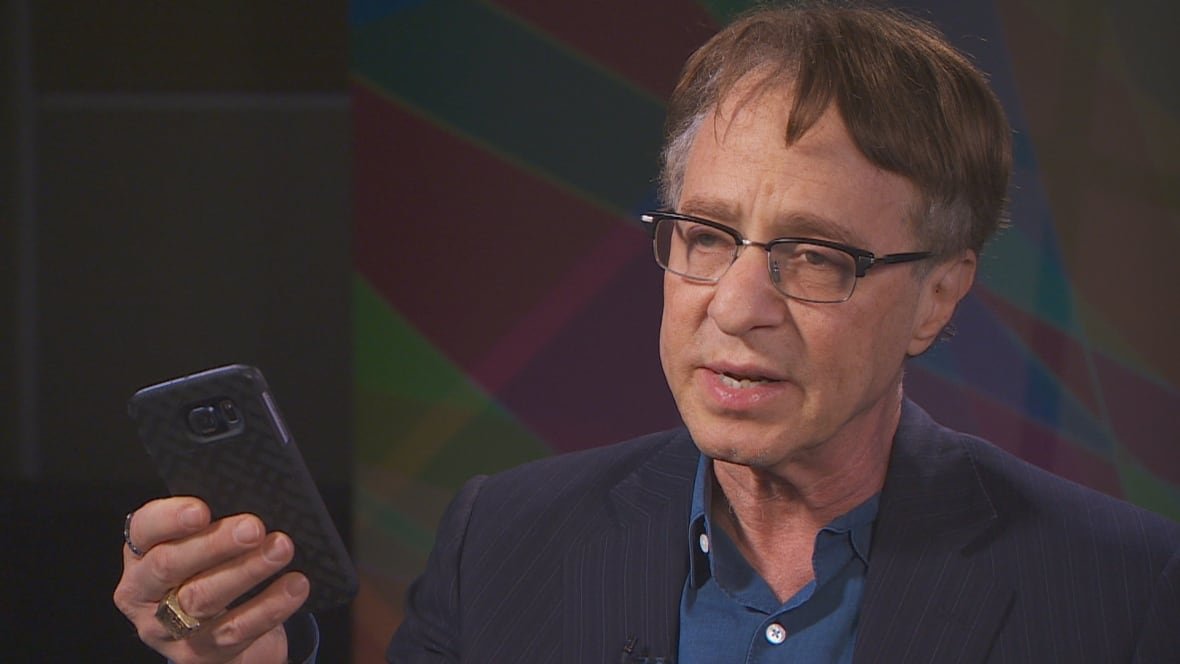 futurist-ray-kurzweil An older man is holding up a cellphone wearing glasses with short hair.