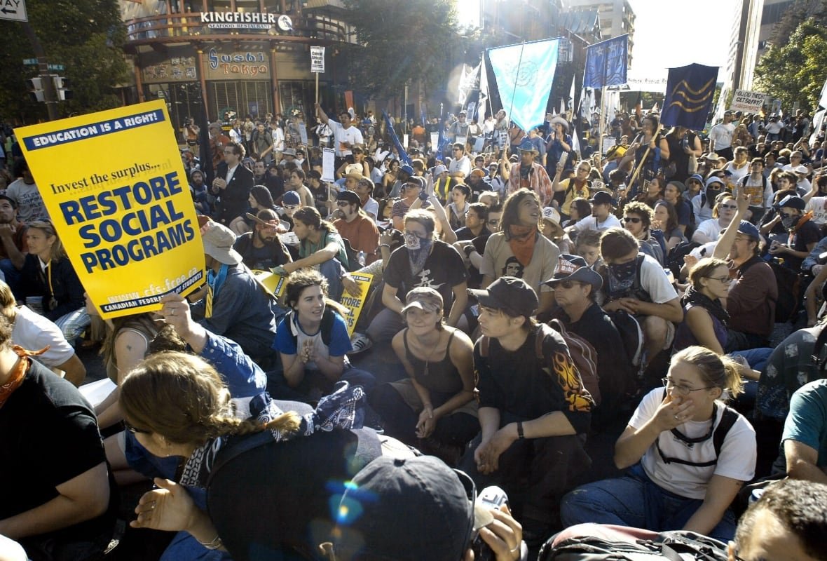 People are pictured in a crowd. A sign reads Restore Social Programs.