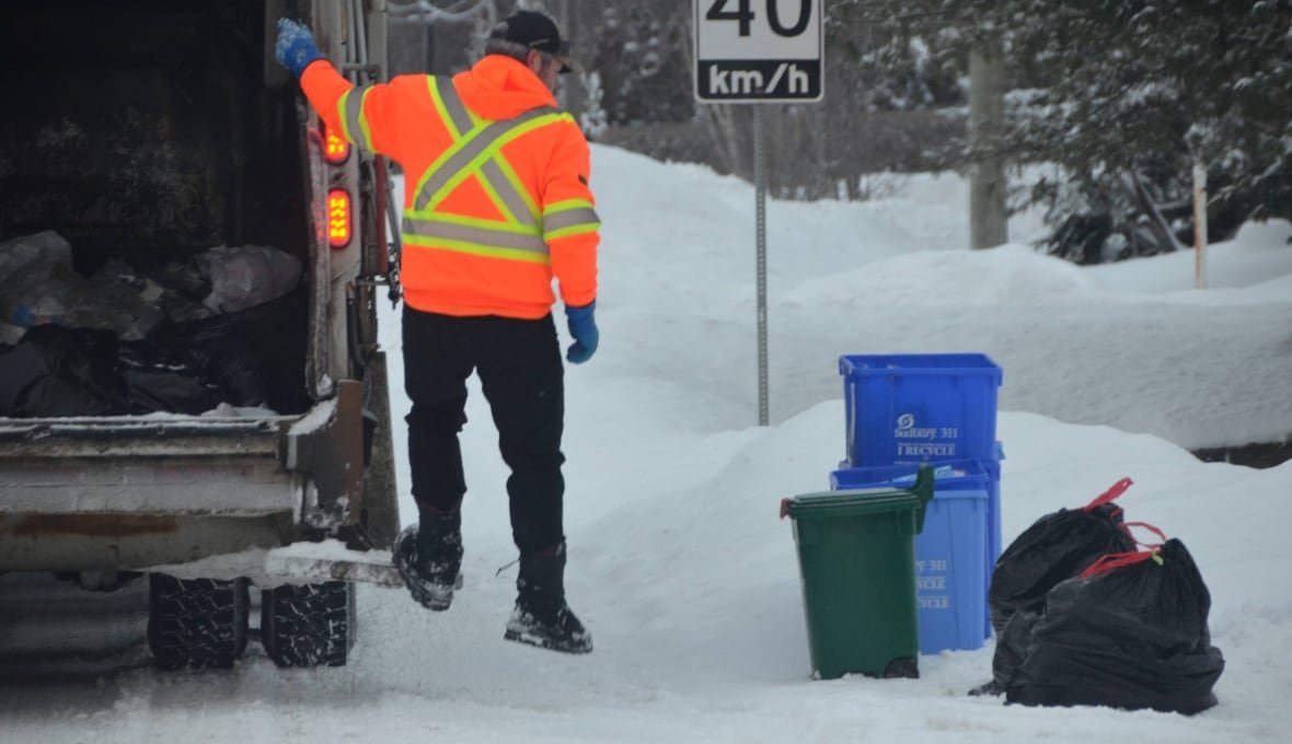 A garbage man hops off a truck and moves toward some bags and bins on a snowy day