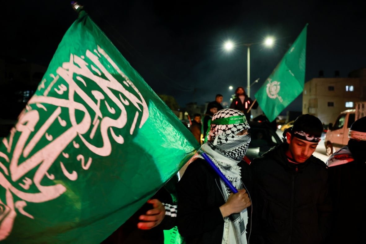 israel-palestinians-gaza-ceasefire-2 A masked man moves through a large crowd at night, carrying a large green flag.