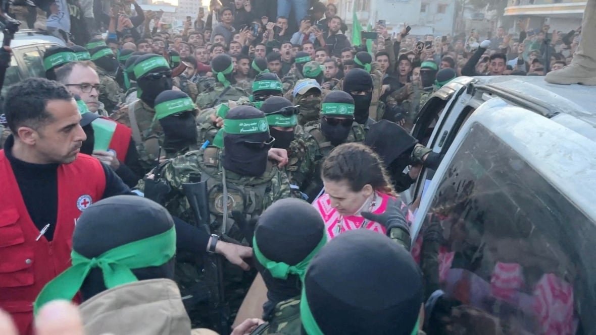Amid a large crowd, a woman is surrounded by masked men holding automatic weapons. Another man, wearing a red vest, reaches out to her.
