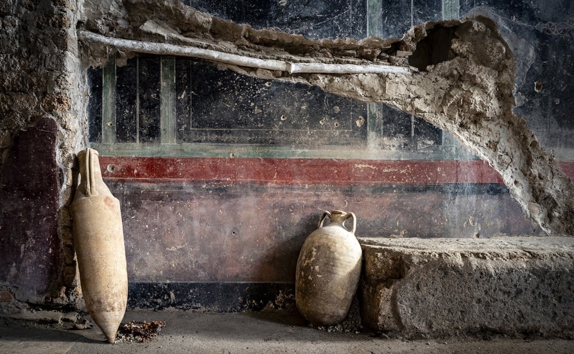 italy-pompeii-1 Two jars lean against a dusty wall covered in smudged red and black stripes for decoration. There is a large chunk of the wall that has been damaged and an oblong shape, perhaps a pipe, runs along the line of the damaged, exposed section.