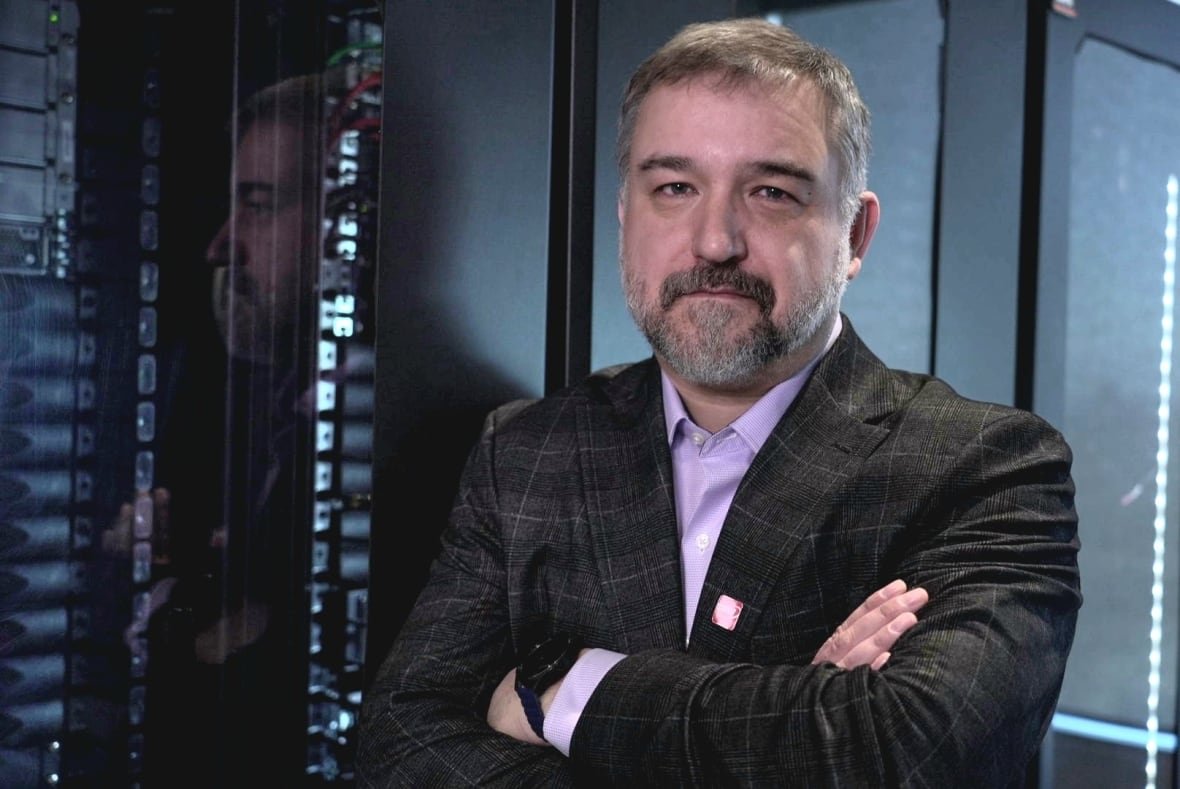 A man in a dark, checked jacket and lavender button-down shirt stands with his arms crossed as he leans against the glass front of a bank of computer servers.