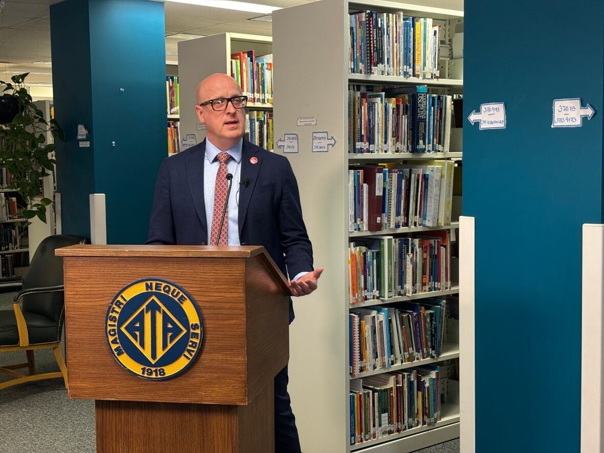 Schilling, wearing a navy blue suit and red tie, stands at a lectern in the library of the Alberta Teachers' Association's Edmonton headquarters. Numerous shelves of books are visible behind him.