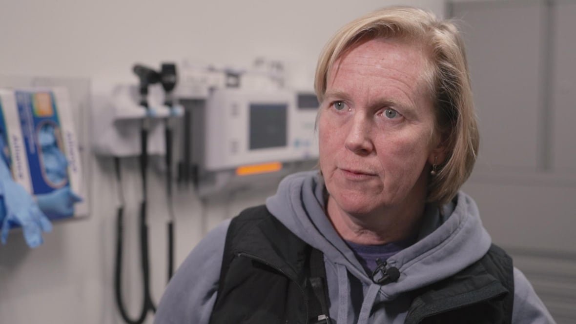 keren-elumir A blonde woman wearing a grey hoodie and a black vest stands next to medical equipment, like sterile gloves and an otoscope.