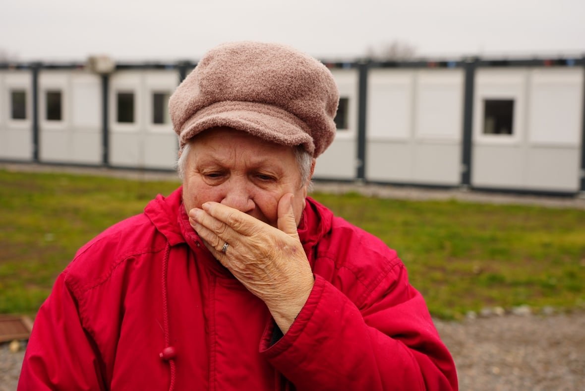 lubov-tymofeeva An older woman in a red coat and beige hat holds a hand to her mouth, looking downward.