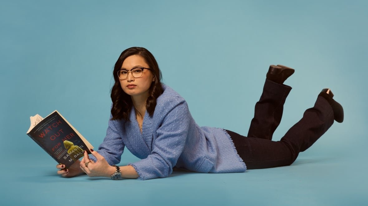 An Asian woman wearing glasses and a blue blazer lies on the floor while holding a book.