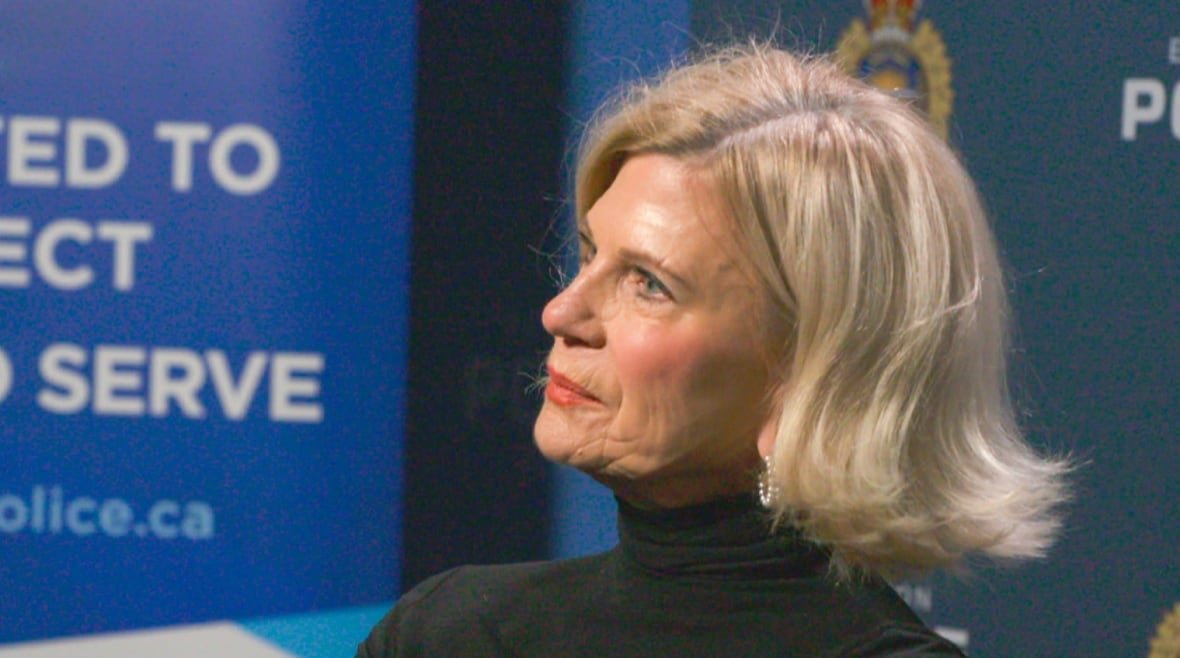 An older white woman, with short blonde hair, is wearing a black turtleneck shirt while sitting in a conference room. Small hoop earrings hang from her earlobes.