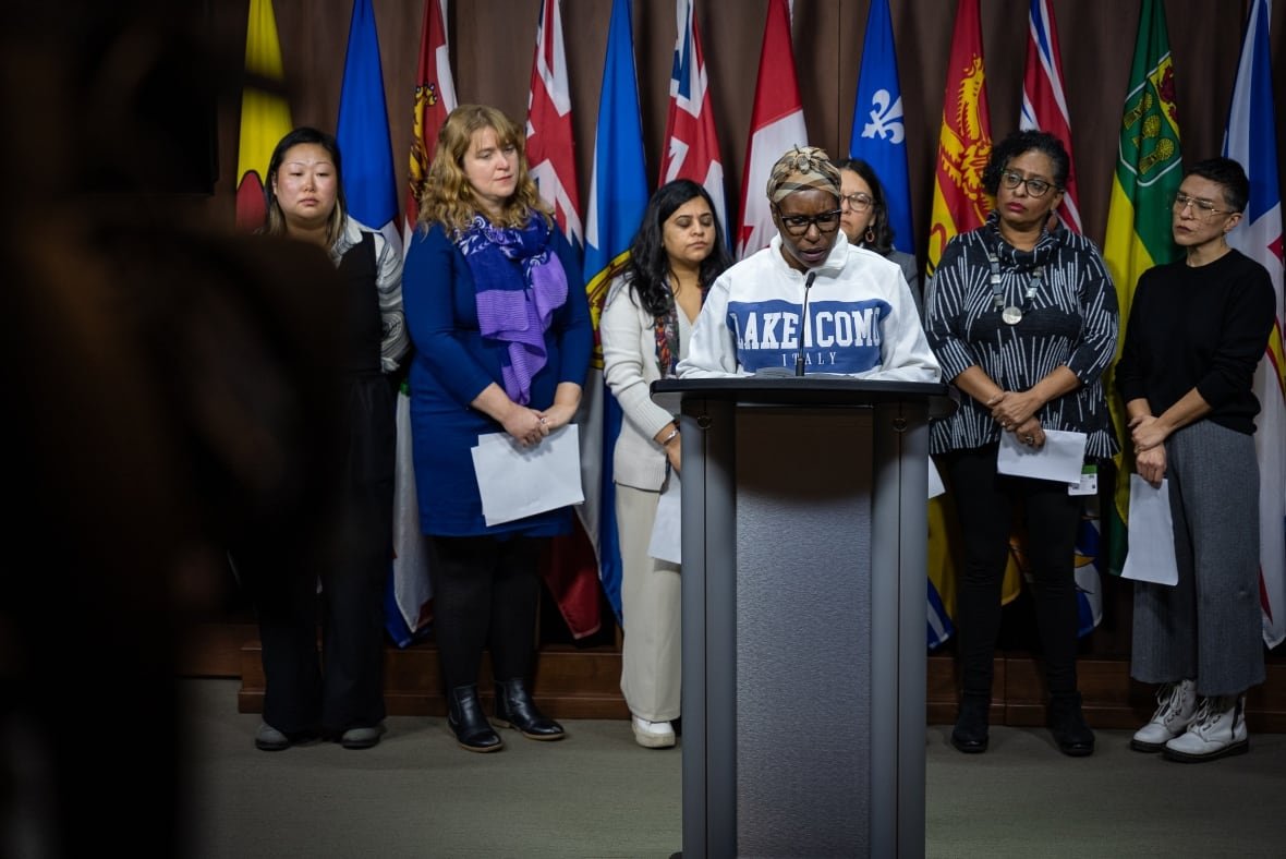 monieya-jess-media-conference A woman wearing a hoodie and a head scarf speaks at a podium flanked by six other women.