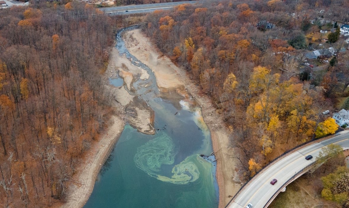 An aerial shot shows a blue-green water source that has clearly shrunken, with brown banks stretching up on either side of the water. It is fall and the surrounding trees have orange leaves or no leaves.