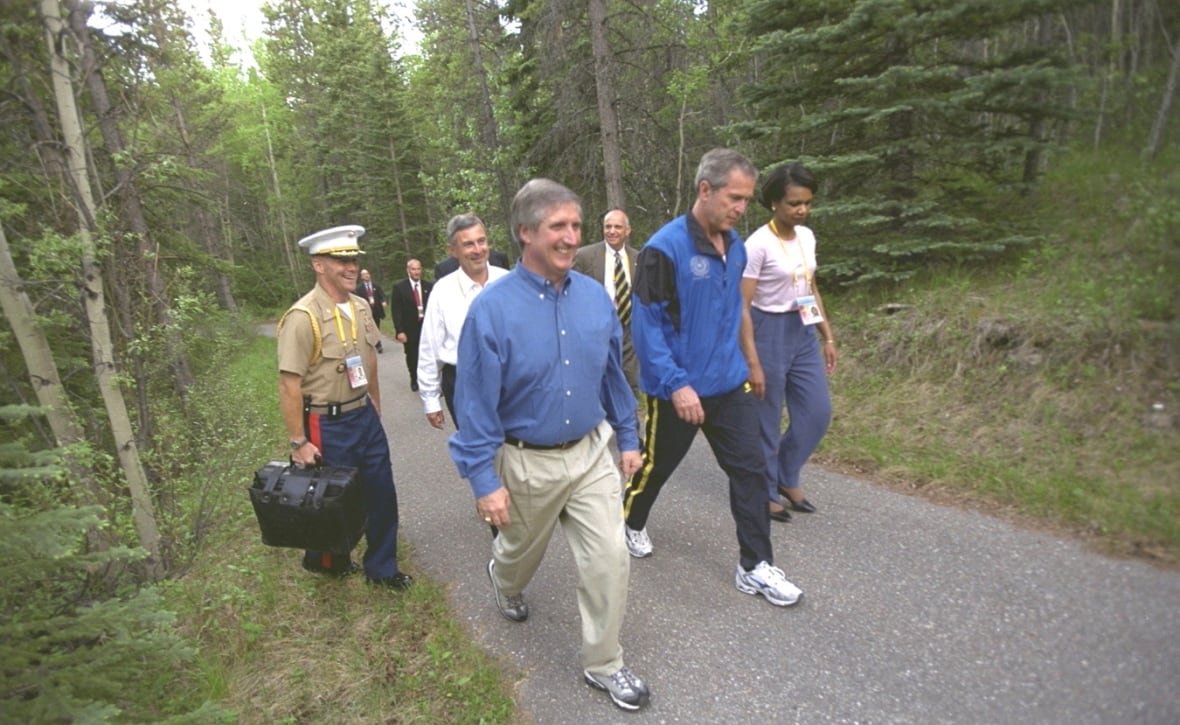 People walk through woods. One individual is holding a satchel.