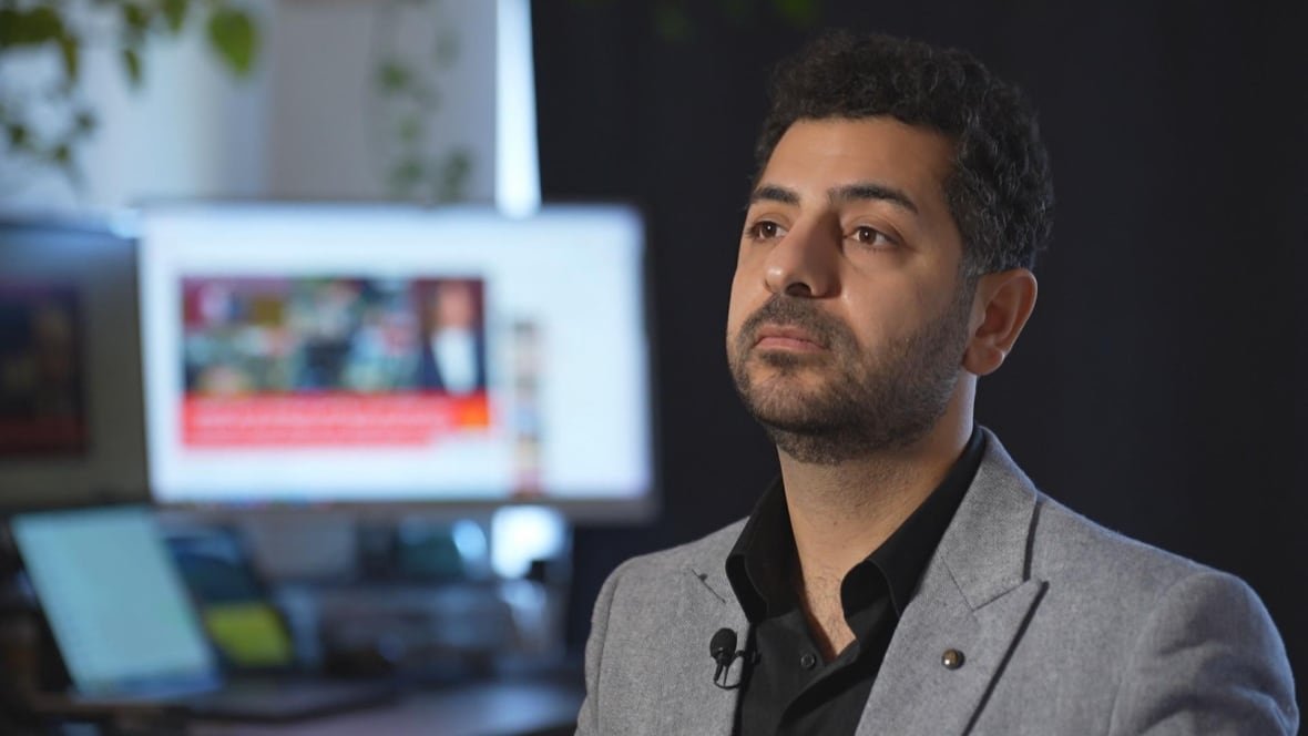 A man wearing a grey suit jacket and black shirt stands in front of computer screens.