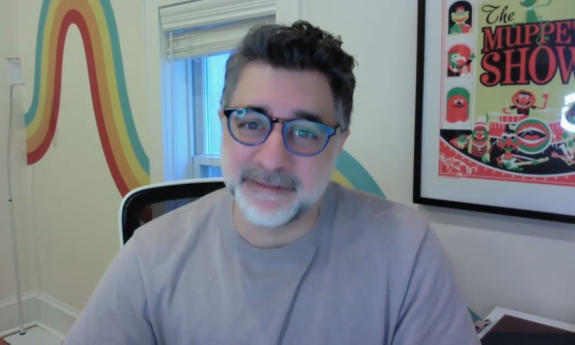 A man in a grey t-shirt, black plastic glasses, with a white beard and darker hair sits looking into a webcam.