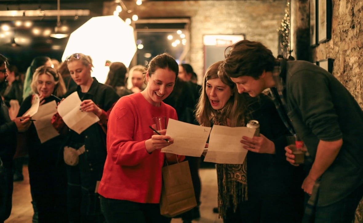 onyx-storm-3 A group of three people crowd around two pieces of paper held by two of them, reacting with amusement at what they are reading. In the background, more people carrying more pieces of paper are visible. They are inside a building with rough brick and warm lighting.