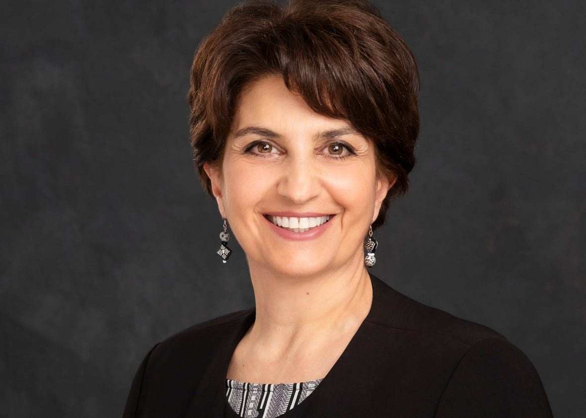 A portrait of a smiling women with short, dark hair and wearing a black jacket and patterned shirt.