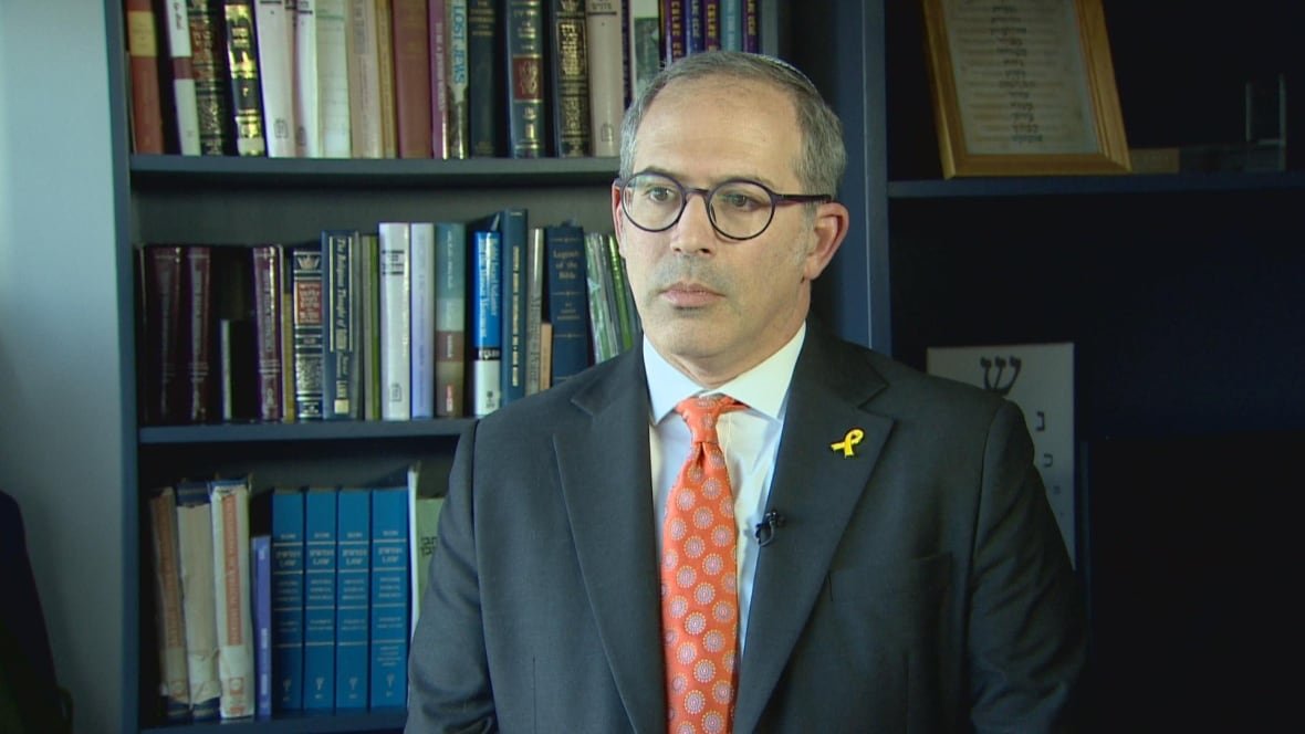 A man with a yellow ribbon pinned to his suit jacket lapel stands in front of a bookcase.