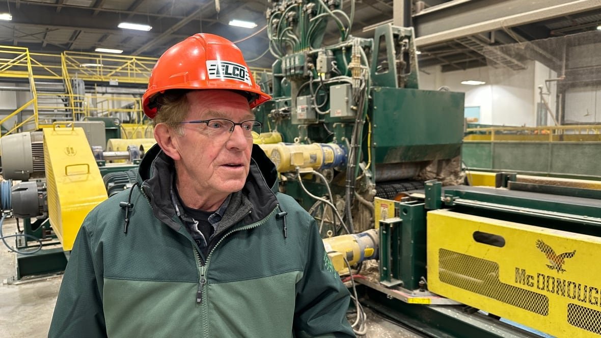 A man in an orange hard hat is shown with heavy machinery at a sawmill shown behind him.