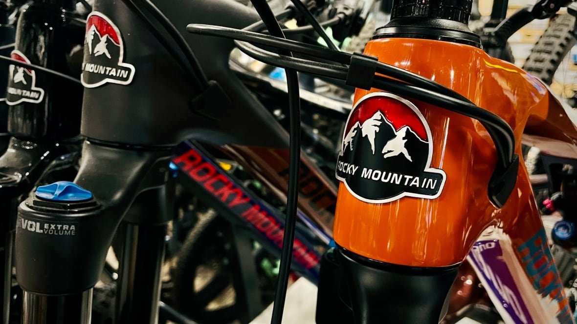 A bright orange mountain bike is pictured with the Rocky Mountain logo to the right of the photo, with two black Rocky Mountain bikes on the left.