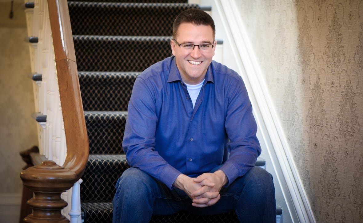 A man sits on a staircase and smiles. He's wearing glasses and a blue collared shirt.
