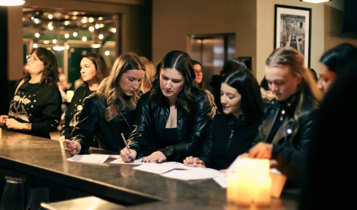 spaniel A group of woman, all wearing black, lean over pieces of paper sitting on a bar. A crowd is visible beyond them, and there is warm lighting throughout.