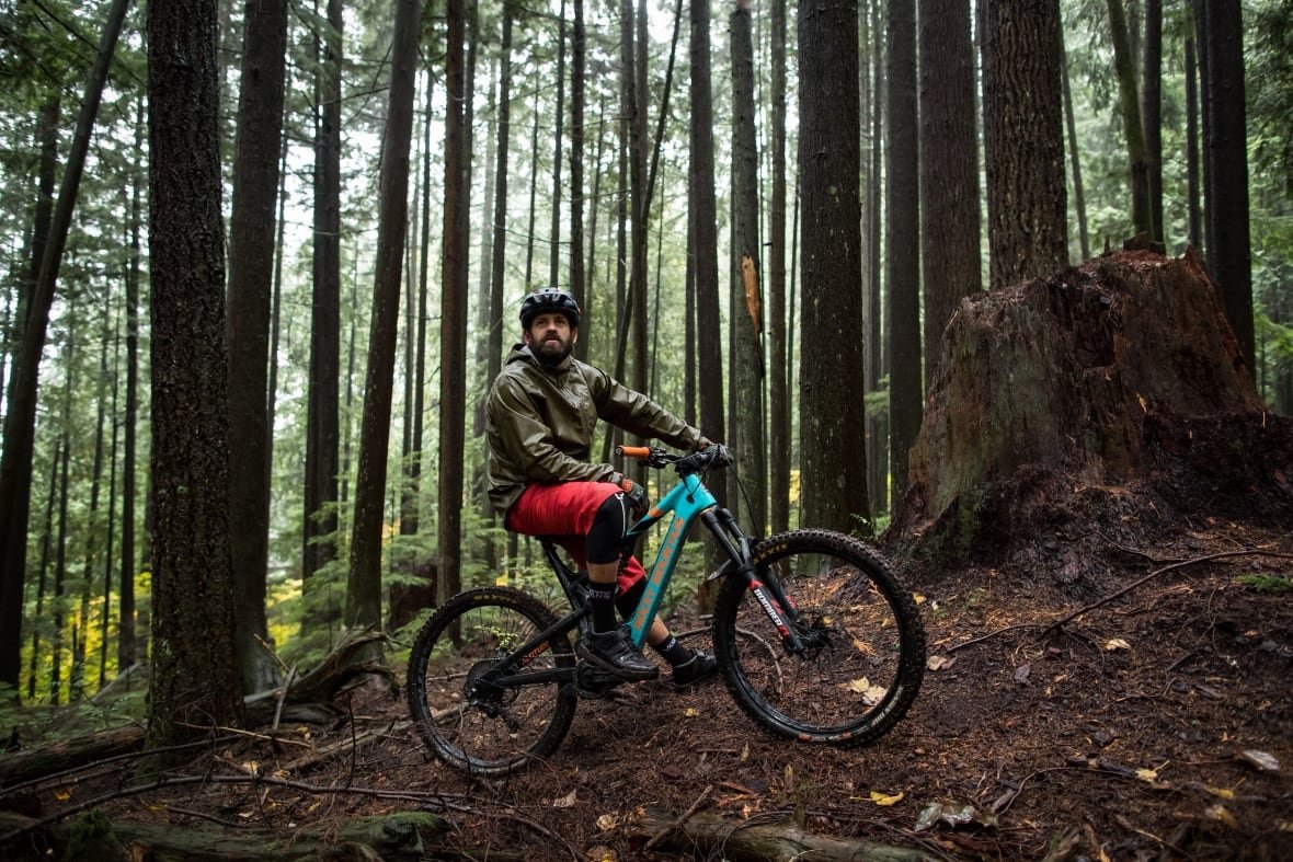 A man in protective mountain bike gear is sitting on a full suspension mountain bike in a temperate rain forest.