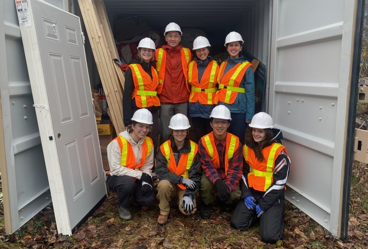 Eight people, all wearing orange safety vests, pose for a photo in two lines, with four standing in the back and four kneeling in front. They appear to be standing between several giant doors.