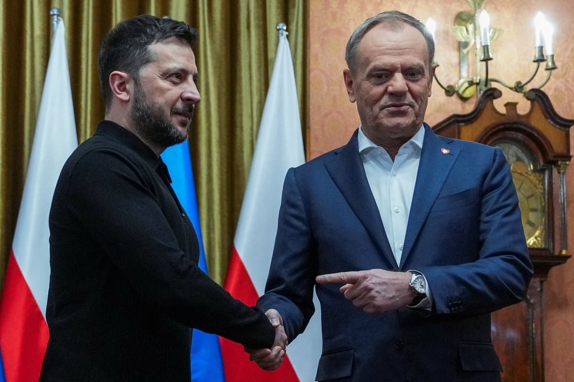 ukraine-crisis-poland Two men, one cleanshaven and wearing a blazer and collared shirt and the other bearded and wearing a sweater, shake hands amid a backdrop of flags.