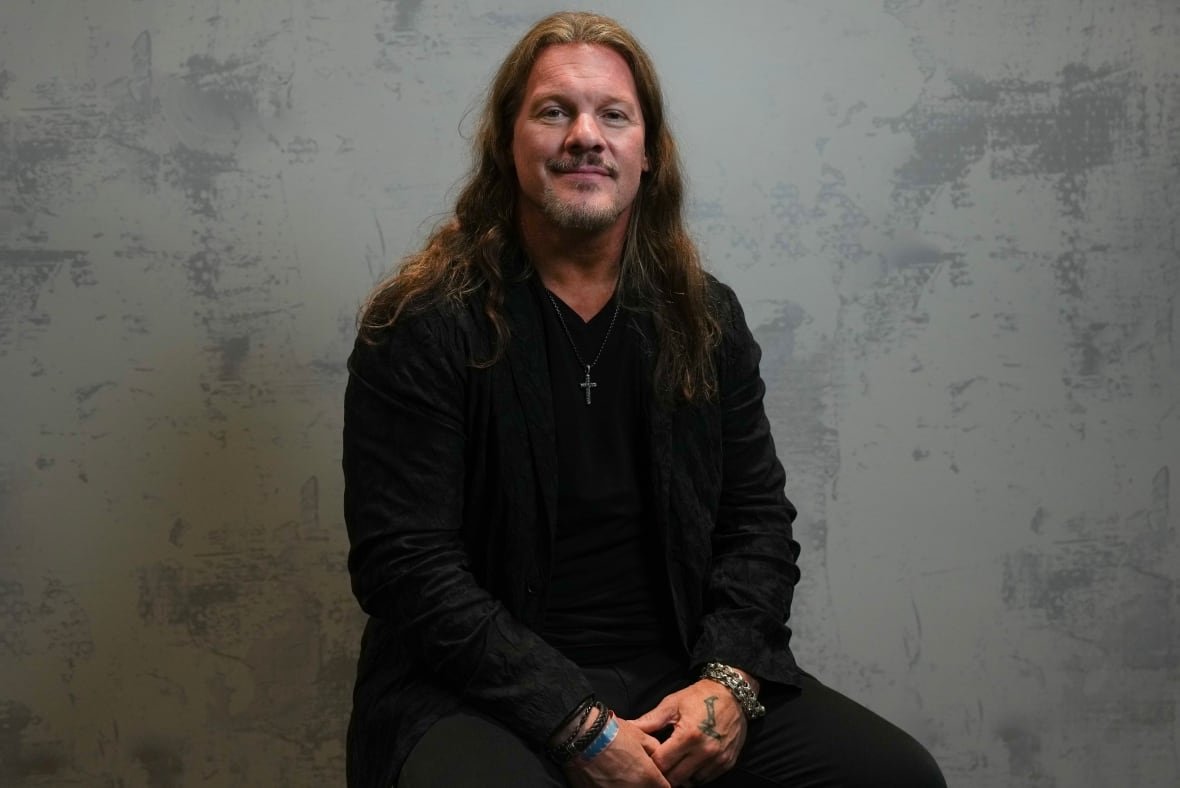 A man with long hair sits on a stool.