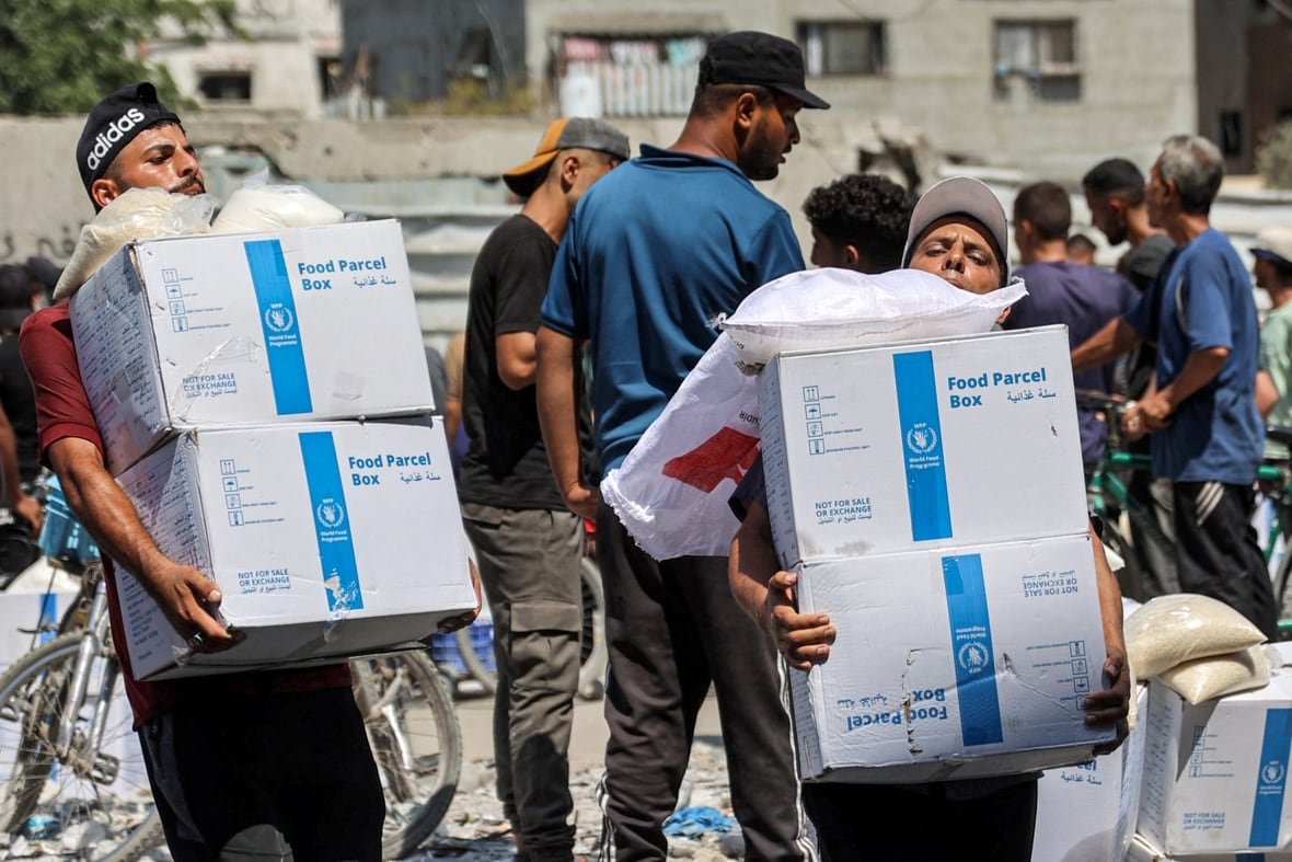 Two men are shown lifting stackes of boxes in an outdoor photo as other people mill around.