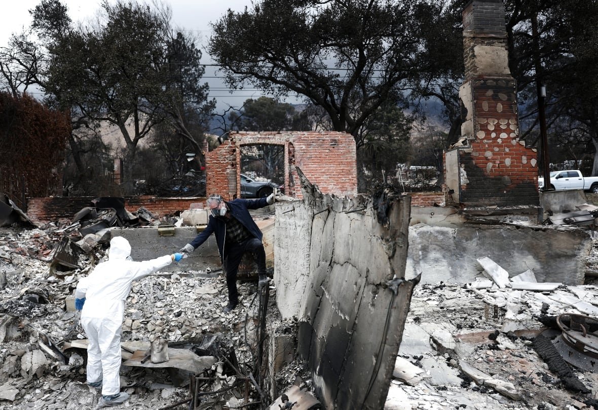 Two people are shown walking among the charred ruins of a heavily destroyed building after a fire.