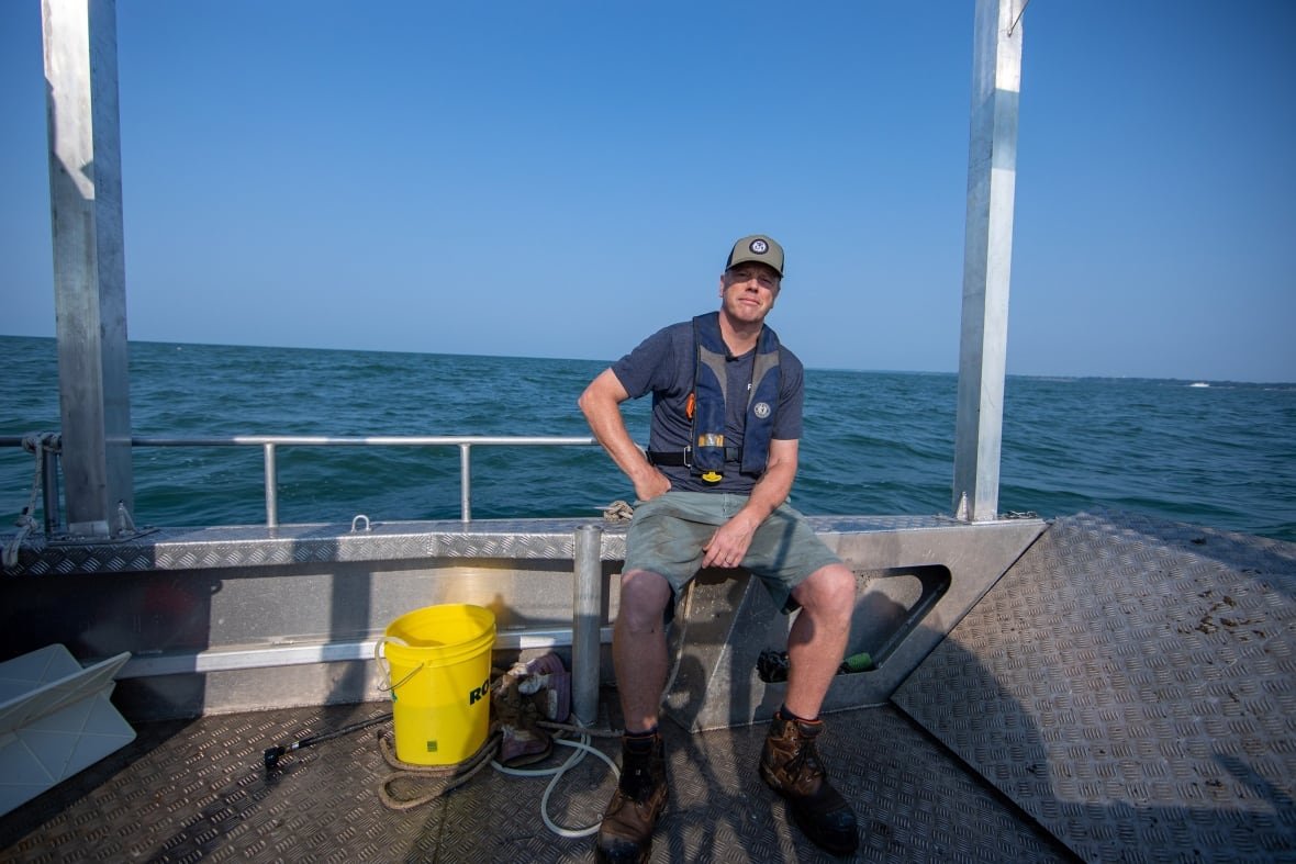 A man wearing a blue cap, shorts and a t-shirt sits on the edge of a small metal research boat. His hand is on his hip and he's slightly grinning. Behind him, blue water meets the horizon. A yellow bucket, rope and various gear sits at his feet. The sun is shining.