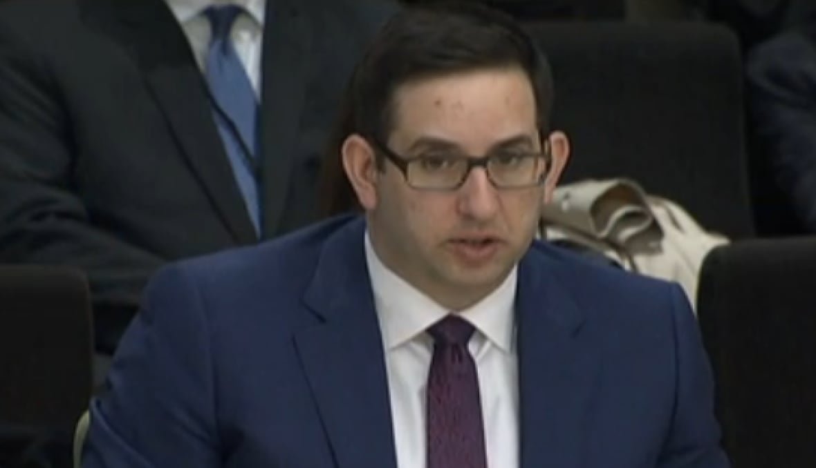 Bespectactled and seated man talks into microphone.