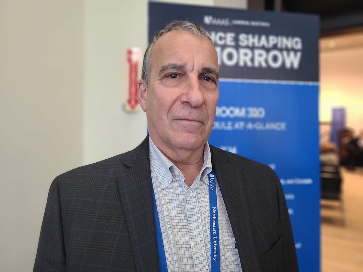 A man with greying hair wearing a blazer and checked button-up shirt looks to the camera. Behind him, a poster reads "science shaping tomorrow."